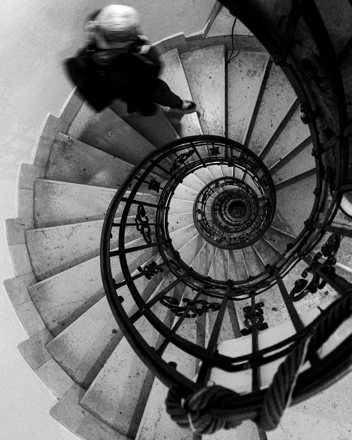 Spiral staircase seen from directly above, its ironwork coiling away to a point, with a figure blurred by movement on the top steps
