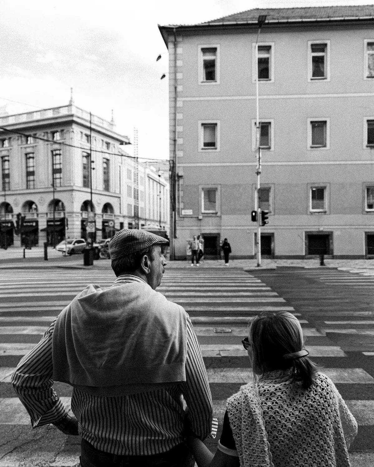 An older couple seen from behind at a pedestrian crossing, a sweater over his shoulders, their hands joined at the bottom of the frame