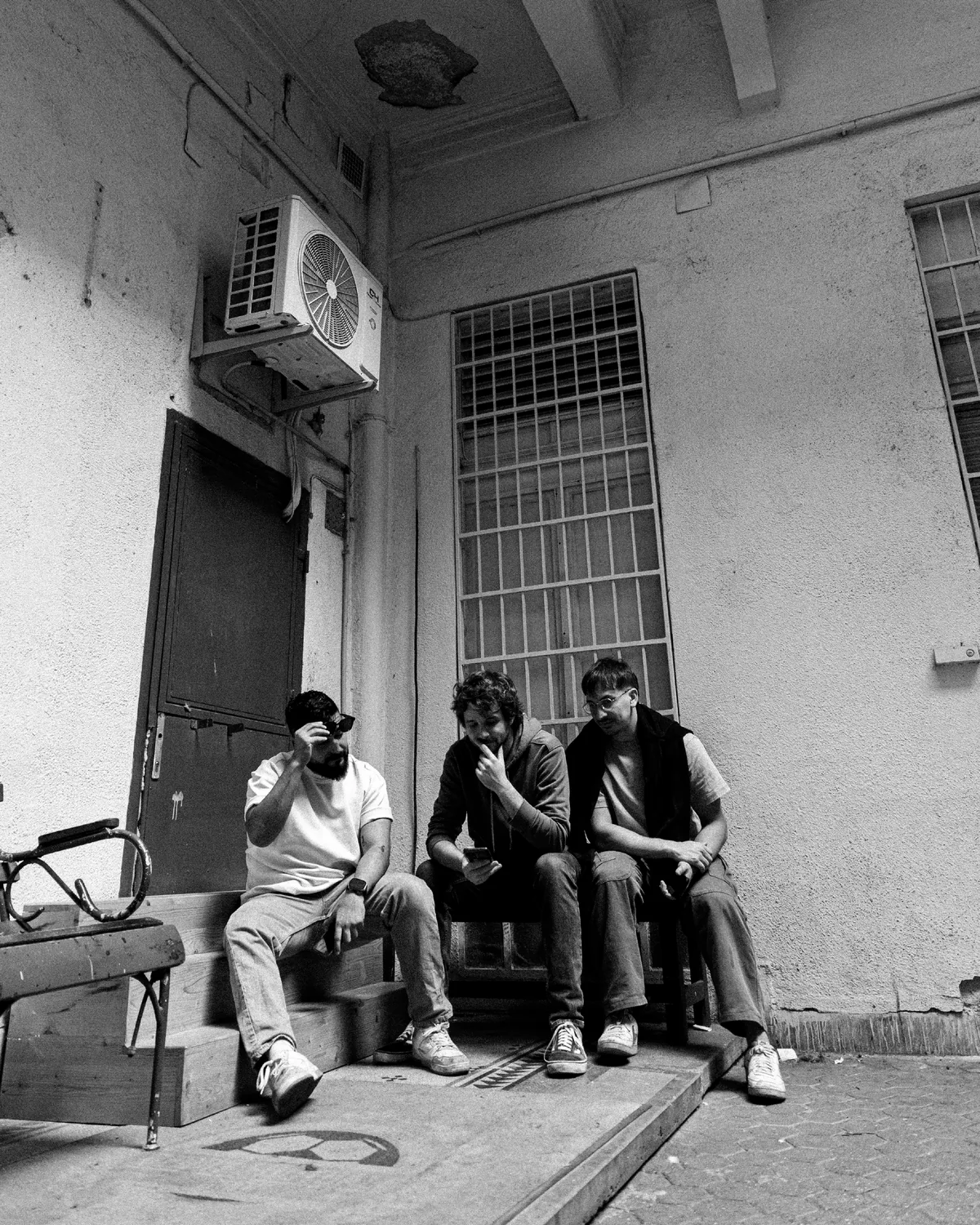 Three men sitting on a step in a courtyard, one shielding his face, one looking down at a phone, one looking away